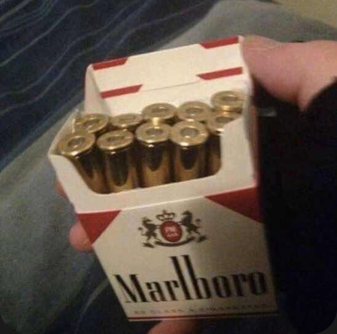 The image displays a person holding a standard red and white Marlboro branded cigarette pack that has been opened. Instead of tobacco cigarettes filling the slots of the pack, brass-colored handgun bullets are placed inside, perfectly sized to replace the cigarettes. The visual gag acts as a dark ironic joke drawing an equivalence between the lethal health consequences of smoking cigarettes and the deadly effect of being shot with a bullet.