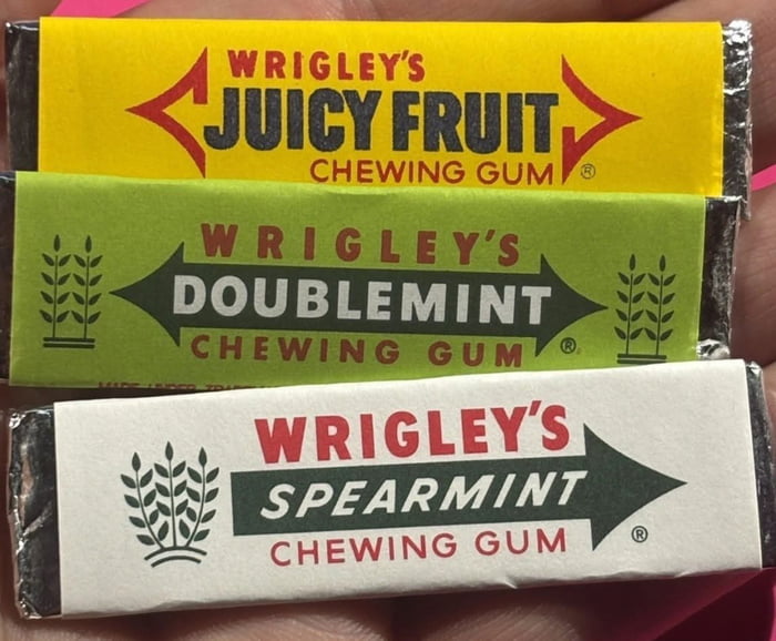 The image presents a close-up shot of three individual Wrigley's chewing gum sticks held in a person's hand, stacked vertically. From top to bottom: 
1. **Juicy Fruit**: A bright yellow wrapper with bold red 'WRIGLEY'S' text, dark blue 'JUICY FRUIT' text, and red arrow graphics pointing inward toward the flavor name, with 'CHEWING GUM' in red below.
2. **Doublemint**: A green wrapper with red 'WRIGLEY'S' text, white 'DOUBLEMINT' text set inside a dark green banner, and stylized wheat plant graphics on either side, with 'CHEWING GUM' in red below.
3. **Spearmint**: A white wrapper with red 'WRIGLEY'S' text, green 'SPEARMINT' text inside a dark green banner (with a right-pointing arrow extension), a stylized wheat plant graphic on the left, and 'CHEWING GUM' in red below.
All three wrappers feature the registered trademark symbol (®) near the bottom right. The background is the skin of a person's hand holding the gum sticks together.
