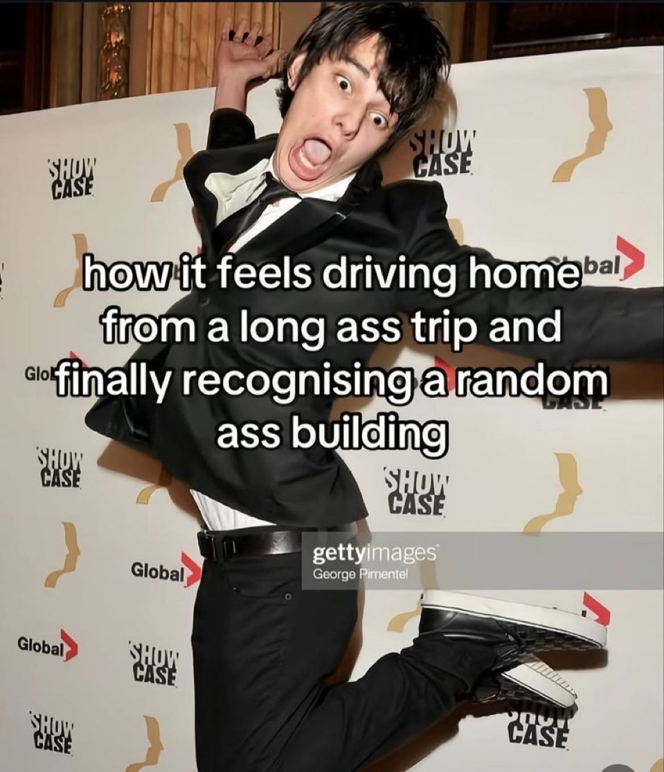 This is a relatable reaction meme featuring actor Devon Bostick (best known for playing Rodrick Heffley in the *Diary of a Wimpy Kid* film series) striking an over-the-top excited, dramatic pose on a red carpet in a black suit. Superimposed text over the image reads "how it feels driving home from a long ass trip and finally recognising a random ass building". The meme humorously captures the exaggerated sense of relief and familiarity people feel when they spot a known local landmark after a long, tiring journey, signaling they are nearly home.