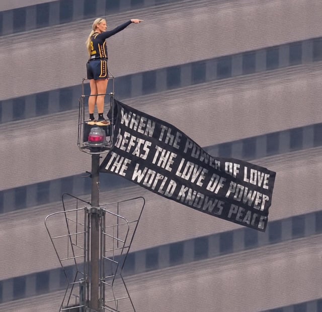 This meme uses a photograph of a female acrobatic performer standing on a platform at the top of an extremely tall utility pole, pointing directly into the distance. A large black banner with a variation of a famous peace quote (with minor typos) hangs from the pole. The comedic premise comes from the title question 'Can you describe the assailant?', which implies this woman, due to her incredibly high vantage point and clear pointing gesture, would be the perfect witness to identify a crime suspect, playing on the absurdity of her elevated position for humorous effect.
