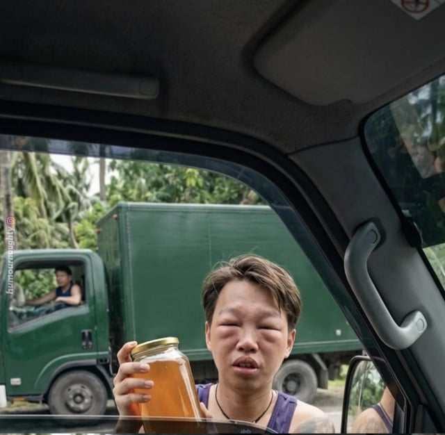 This photo taken from inside a car shows a person holding up a large glass jar of honey toward the camera. The person has extremely swollen, puffy eyes that are clearly irritated from multiple bee stings. In the background, a second man drives a green delivery truck alongside the car. The meme uses a visual pun: the swollen eyes from bee stings serve as 'proof' that this is 100% natural, wild-harvested honey, since the person was stung while collecting it, creating a painful but funny joke.