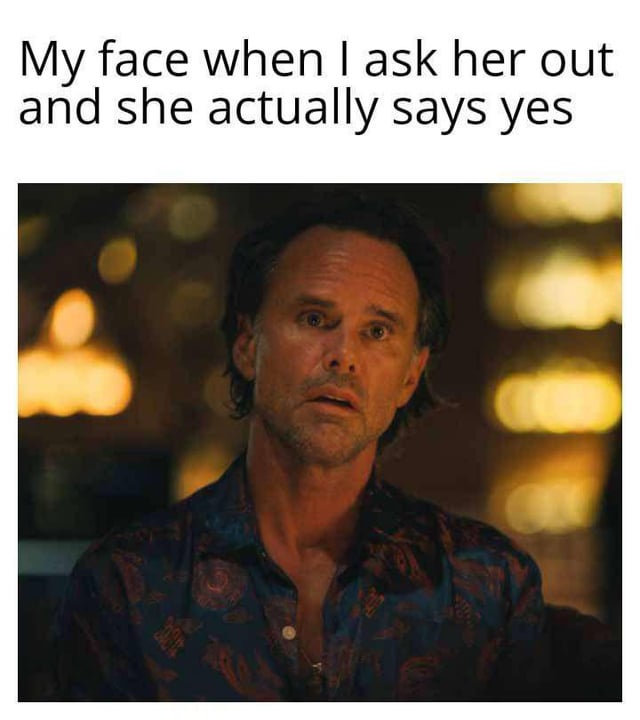 A meme consisting of a top text caption and a screenshot of a man with a shocked, wide-eyed expression. The top text reads: 'My face when I ask her out and she actually says yes'. The man in the image has short, slightly messy hair, a beard stubble, and is wearing a dark blue shirt with a red and gold pattern. The background features blurred warm yellow lights, creating a cozy, indoor setting. The man's expression conveys surprise and disbelief, aligning with the caption's context of unexpected positive response to a date request.