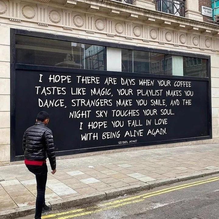 This photograph captures a man standing on a paved city sidewalk next to a street curb, looking at a large hand-painted message on a blacked-out vacant storefront window of an ornate urban building. The message is written in white hand lettering, and shares gentle, uplifting positive wishes for the viewer: it hopes the reader experiences days where their coffee tastes magical, their music playlist makes them want to dance, strangers bring them an unexpected smile, the night sky touches their soul, and ultimately that they fall back in love with being alive. The art is credited to 'Seconds Apart' at the bottom of the message.