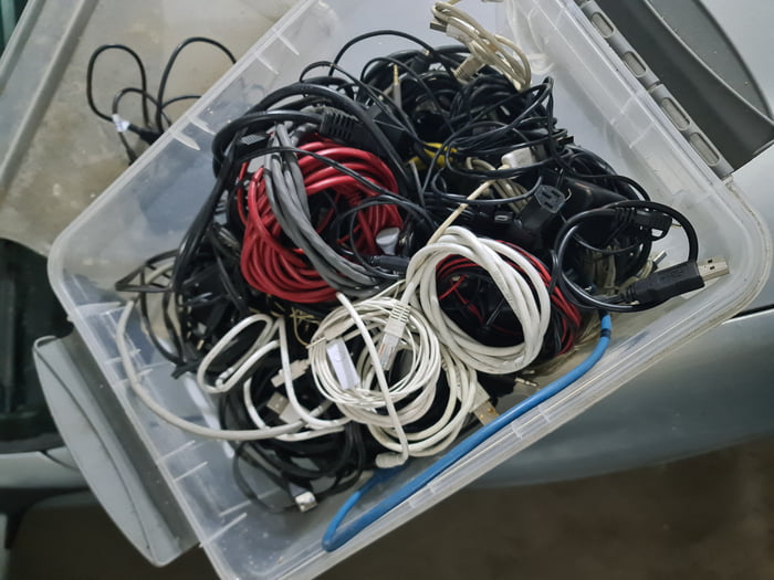 The image displays a transparent plastic storage tote completely filled with a large, disorganized, tangled pile of assorted cables of varying types and colors. Visible cables include USB charging cords, Ethernet network cables, power supply cords, audio cables, and other data transfer cables in black, red, white, grey, and blue hues. The meme plays on the widely relatable trope that many people, stereotypically men, keep a dedicated box full of old, unused cables hoarded over years just in case they might need one of them in the future, and the poster is humorously announcing they finally had reason to use one of the cables from this box.