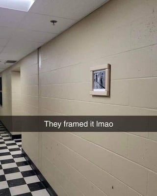 The image captures a long institutional hallway with beige paneled walls and a black-and-white checkered tile floor. A small, neutrally colored picture frame is mounted on the wall as if it is formal, curated artwork, but the image inside the frame is a widely recognizable internet meme. White overlaid text on a dark gray banner across the lower middle of the photo reads 'They framed it lmao', emphasizing the absurd, humorous premise of treating a casual viral internet meme as a respectable piece of wall art.