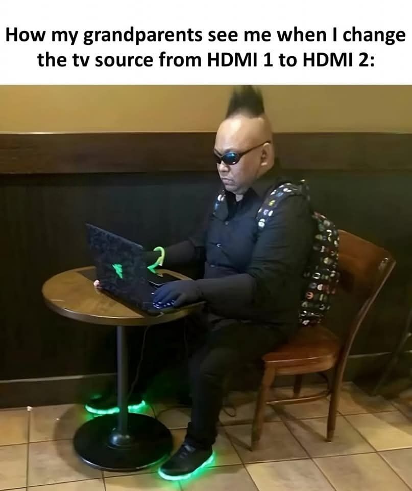 This is a relatable humor image macro. The top of the image has text reading 'How my grandparents see me when I change the tv source from HDMI 1 to HDMI 2:'. Below the text is a photograph of a man with a tall mohawk hairstyle, dark sunglasses, all-black outfit, fingerless gloves, and shoes with glowing neon green soles, sitting at a small round table typing on a Razer brand gaming laptop. The joke exaggerates the common experience where older generations like grandparents vastly overestimate the technical expertise of younger people for extremely simple, routine tech tasks, framing the trivial action of switching a TV's input source as the work of a skilled, edgy elite hacker.