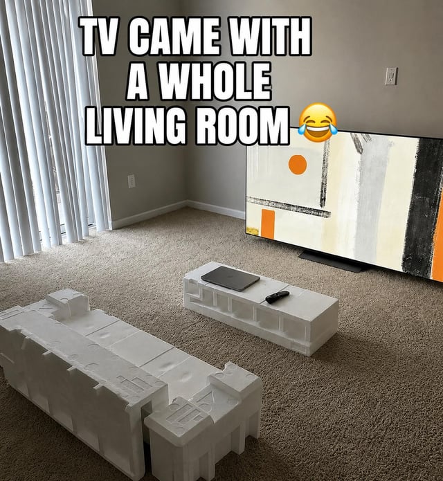 The meme is a photo of an empty carpeted living room with a large new flat-screen TV placed against the wall. The styrofoam protective packaging that came with the TV is intentionally arranged to look like a complete living room set, including a couch, armchair, and coffee table. A laptop and TV remote are placed on the styrofoam coffee table to sell the joke. Overlaid text at the top reads 'TV CAME WITH A WHOLE LIVING ROOM' next to a laughing crying emoji, making a humorous relatable joke about people who buy new electronics before having purchased actual furniture for their home, using the free packaging as temporary furniture.
