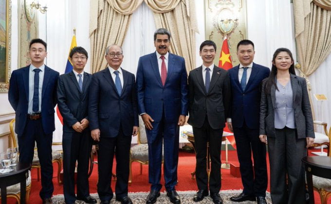 A formal group photograph depicting Chinese officials and Venezuelan President Nicolás Maduro during a diplomatic meeting. The individuals are standing in a line in an ornately decorated indoor setting featuring draped cream curtains, red carpet, and flags (including Venezuela's flag) in the background. All participants are dressed in formal suits, with President Maduro positioned centrally, suggesting a high-level official gathering.