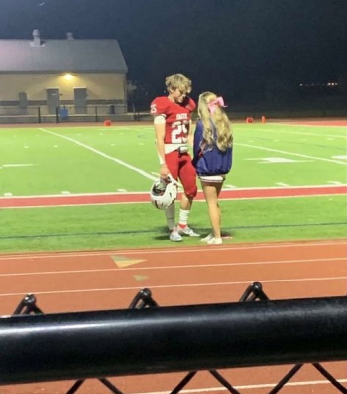 This is a candid night-time photograph taken from spectator stands behind a metal railing, overlooking a high school American football field and adjacent running track. At the center of the frame, a teenage male football player in a red number 25 uniform holds his white football helmet as he stands facing a blonde teenage cheerleader with a large pink bow in her hair, who is wearing a navy cheer jacket and white shorts. The pair are looking at each other in a soft, intimate moment, likely occurring after a football game. The associated meme title communicates that this kind of innocent, nostalgic youthful connection is a priceless feeling that cannot be purchased even with lottery winnings.