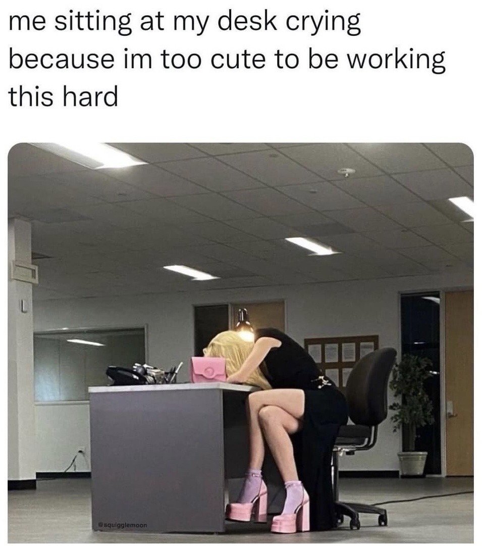 This is a relatable humorous work meme. The top of the image has text stating 'me sitting at my desk crying because im too cute to be working this hard'. Below the text is a photograph of a blonde woman wearing a black dress, light purple socks, and pink platform high heels, slumping face-down over an office desk in an empty, brightly lit corporate office space, appearing visibly upset or frustrated. The joke leans into dramatic, playful self-aggrandizement as a silly excuse for being burnt out from work.