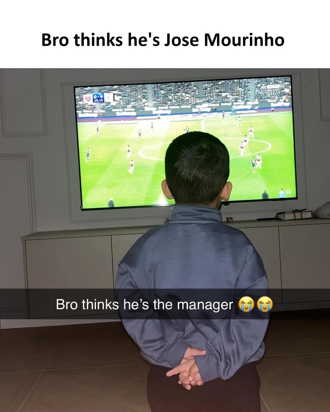 The image shows a young boy photographed from behind, standing with his hands clasped behind his back as he intently watches a professional football match playing on a large wall-mounted television. Text at the top of the meme reads "Bro thinks he's Jose Mourinho", and overlay text at the bottom of the photo says "Bro thinks he's the manager" accompanied by two crying laughing emojis. The joke comes from the fact that famous professional football manager Jose Mourinho is famously often photographed standing on the match touchline with his hands held behind his back while observing games, so the boy's identical posture makes it look like he is acting as the manager of the team playing on TV.