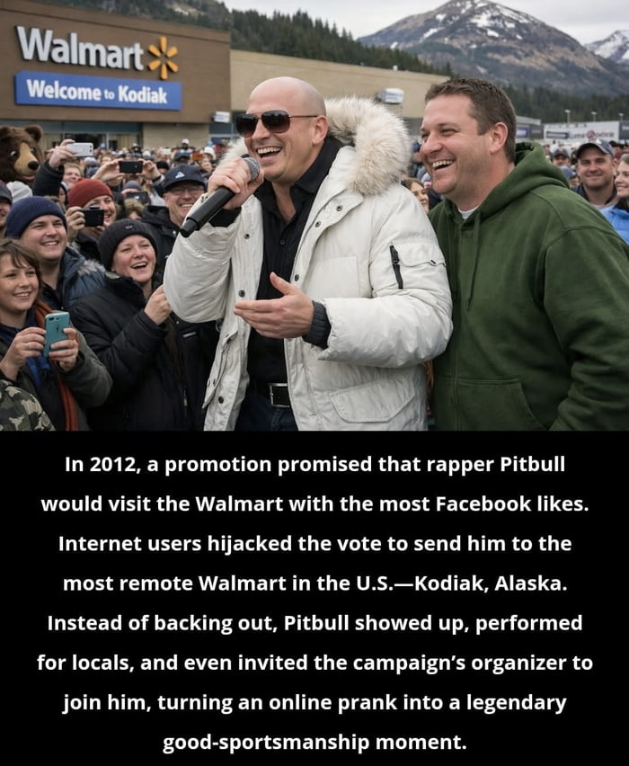A photograph capturing rapper Pitbull performing in front of a crowd outside a Walmart in Kodiak, Alaska. Pitbull, wearing a white jacket with a fur-lined hood, holds a microphone and smiles broadly, while a man in a green hoodie stands beside him, also smiling. The background shows the Walmart storefront with a sign reading 'Welcome to Kodiak' and a mountainous landscape. The scene reflects the 2012 event where internet users voted to send Pitbull to the remote Alaskan Walmart, and he honored the commitment with a local performance.