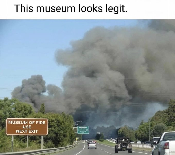 The image displays a multi-lane highway scene on a clear day. A brown roadside exit sign on the left reads 'MUSEUM OF FIRE USE NEXT EXIT'. Directly in the distance past the indicated exit, an enormous thick cloud of dark black and grey smoke rises high into the sky, showing a large active fire is burning at that location. The top of the meme has sarcastic text 'This museum looks legit.' which jokes about the coincidence that the exit for the Museum of Fire appears to lead directly to a massive real fire, ironically making the museum seem to literally match its name by being engulfed in flames.