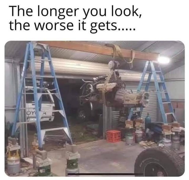 The image displays an extremely unsafe, jerry-rigged setup inside a garage workshop used to hoist a heavy car engine. Two A-frame ladders are positioned on either side, each balanced precariously on top of stacked unstable objects including gas canisters, plastic bottles, and wooden blocks instead of being placed on flat, stable ground. A long rough wooden plank is laid across the tops of the two ladders, and the car engine is suspended from this plank via straps and a winch. Text overlaid at the top of the image reads "The longer you look, the worse it gets.....", pointing out that multiple layers of OSHA (Occupational Safety and Health Administration) safety violations become apparent the more you examine the setup. A partially visible Toyota truck, red plastic crate, tires, and assorted workshop tools are also visible in the background of the garage space.