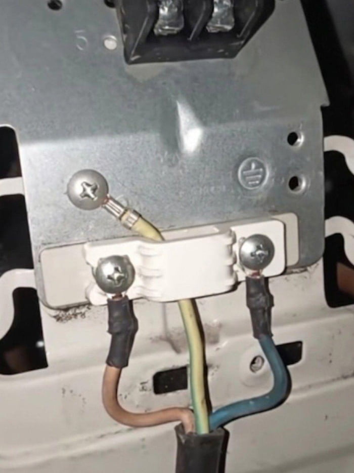 The image displays a close-up of botched internal electrical wiring on a metal equipment chassis. The yellow-green striped ground wire, which is required to be secured to the clearly marked grounding point (the stamped earth symbol visible on the silver metal plate) for safety, is incorrectly connected to the center terminal of the white plastic wiring connector, between the brown live and blue neutral wires. This is a severely unsafe electrical installation that poses extreme electrocution and fire risks, prompting an instinctive 'oof' reaction from viewers who recognize the dangerous amateur mistake.