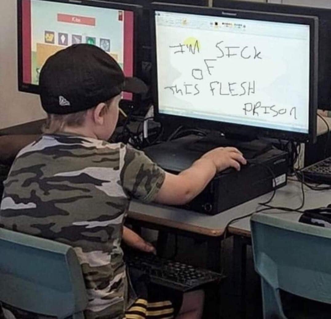 The image captures a young toddler boy from behind, wearing a backwards black baseball cap and a camouflage print short-sleeve t-shirt, seated at a desk in front of two desktop monitors. The left monitor displays a colorful children's game interface, while the right monitor shows messy, childlike handwritten text that reads 'I'M SICK OF THIS FLESH PRISON'. The comedic effect arises from the extreme juxtaposition between the subject, a small child who would typically be expected to use the computer for games or simple activities, and the deeply dark, existential, edgy sentiment of the text, which is usually associated with online edgy humor, occult themes, or adult burnout.