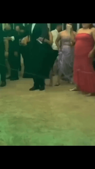 This low-resolution candid photo captures a group of people at a formal event, likely a prom, wedding, or gala, evidenced by attendees in tuxedos and formal evening gowns standing on a carpeted event floor. A forced perspective optical illusion forms the core comedic gag: the bent lower leg and shoe of a person standing behind a man in a black formal suit align perfectly with the suited man's feet, making it appear as if the man is casually standing on a skateboard at the fancy event, matching the joke title of 'Real Pro Skater'. All attendees' heads are cut off by the top edge of the frame.