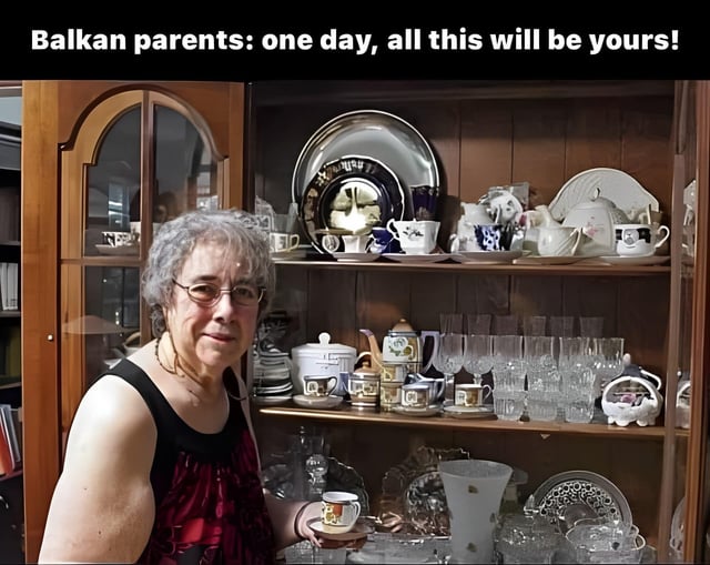 This edited absurdist meme features an older woman's head photoshopped onto a muscular man's shoulder and arm, as she stands in front of a large glass-fronted wooden china cabinet completely packed with an extensive collection of porcelain teacups, plates, glasses, and dishware. The text at the top of the image reads "Balkan parents: one day, all this will be yours!". The joke plays on a common cultural stereotype that Balkan parents and grandparents accumulate massive collections of porcelain and dishware to pass down as inheritance to their children; the edited muscular arm implies the inheritor needs enormous strength to carry and handle this huge, heavy collection of dishware.