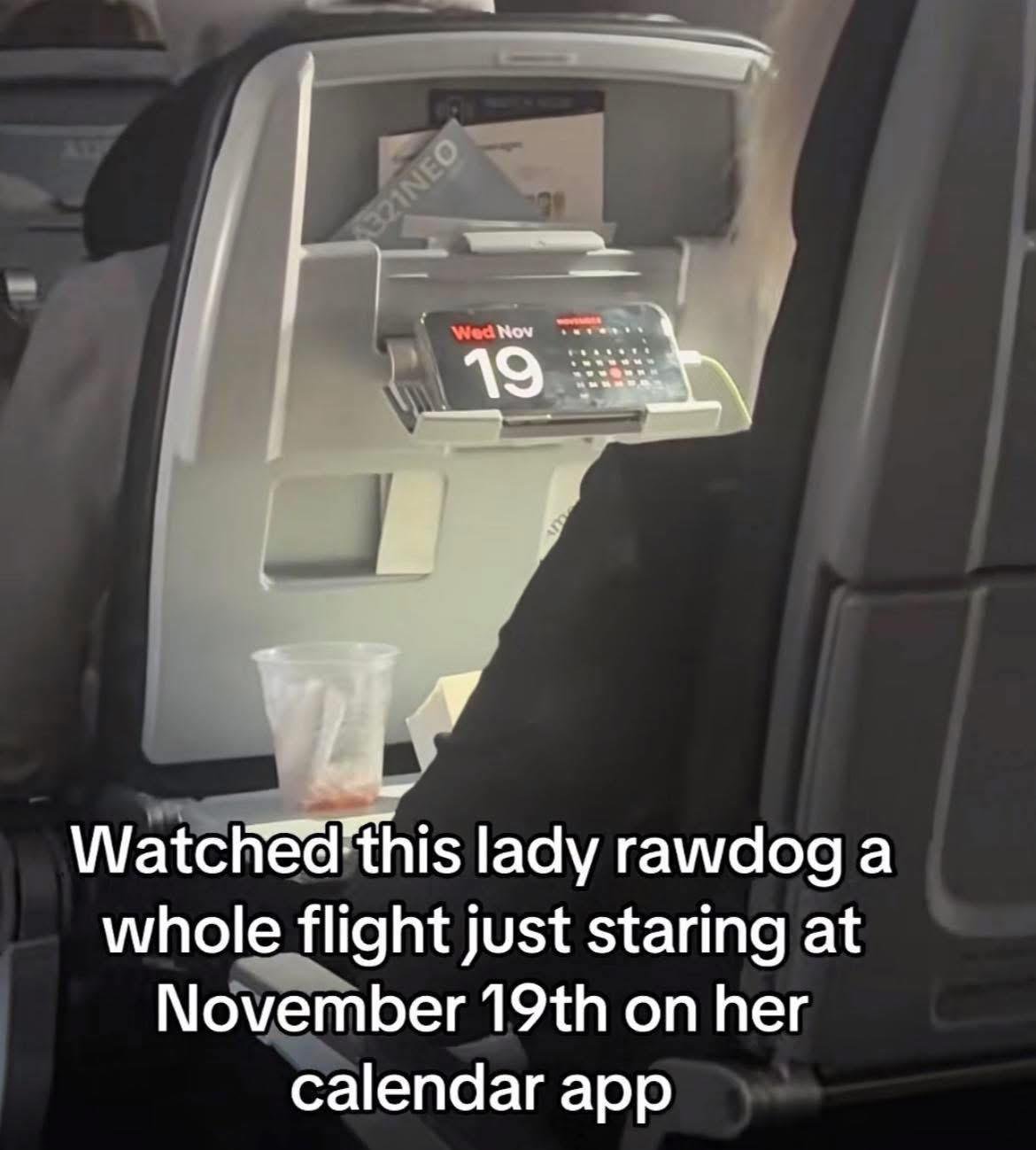 A photograph taken from an airplane seat showing the back of the seat in front, which has a calendar app displaying 'Wed Nov 19'. There's a plastic cup with liquid in the seat pocket. The caption at the bottom reads: 'Watched this lady rawdog a whole flight just staring at November 19th on her calendar app'.