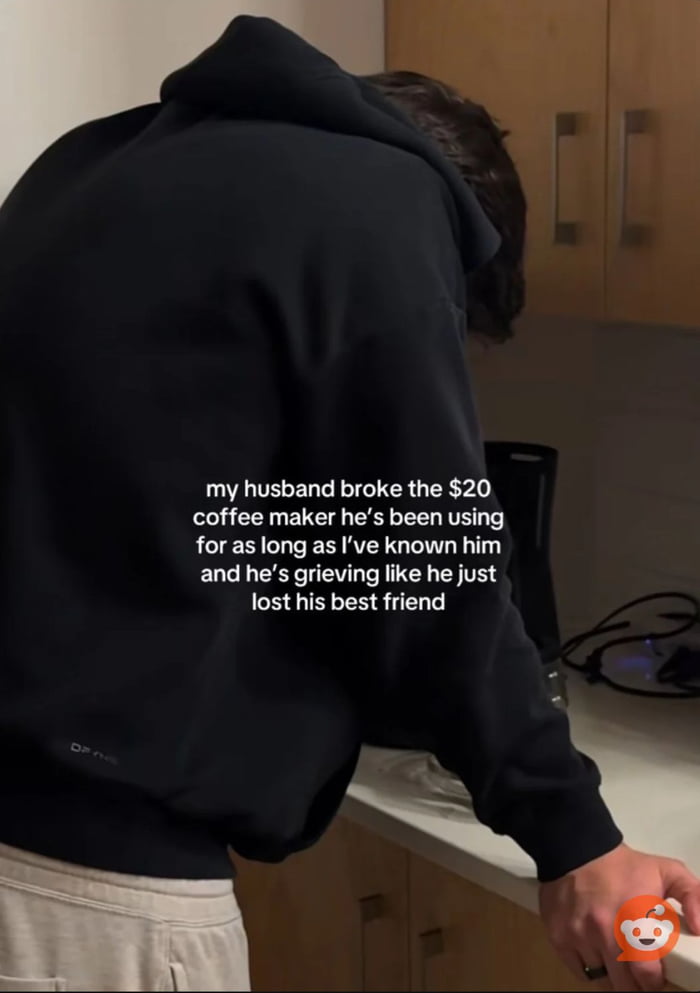 A photograph capturing a man in a black hoodie leaning over a kitchen counter with his head低垂, appearing deeply distraught. The scene is set in a domestic kitchen with wooden cabinets and a coffee maker visible on the counter. Overlaid text explains the context: the man (referred to as 'my husband') has broken a $20 coffee maker he has used for many years, and his grief is humorously exaggerated to resemble mourning the loss of a close friend. The image blends everyday domesticity with relatable humor, highlighting the unexpected emotional attachment to mundane objects.