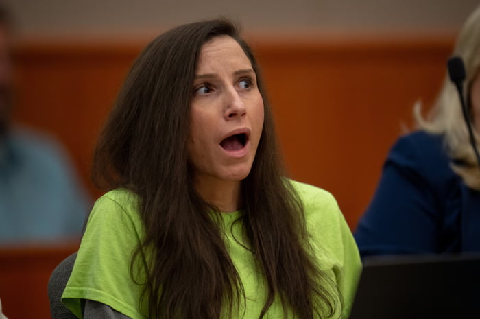 The image captures a real woman with long dark brown hair, wearing a bright lime green short-sleeved t-shirt over a grey long-sleeve top, sitting in a courtroom setting. She has an expression of extreme shock and disbelief, with her mouth wide open and eyes wide, looking up towards her right. The background features blurred wooden paneling typical of a courtroom interior, with partially visible other people seated nearby. The photo is taken in the moment the woman hears that she has been sentenced to life in prison.