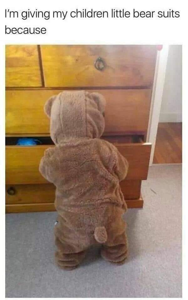 This is a lighthearted wholesome pun meme. The top text reads 'I'm giving my children little bear suits because', below this text is a photo of a toddler (viewed from behind) wearing a full fuzzy brown bear onesie standing in front of a wooden dresser with one open, empty drawer. The joke relies on the homophone pun: 'bear suit' sounds exactly identical to 'bare drawer', referring to the open empty (bare) drawer the toddler is interacting with.