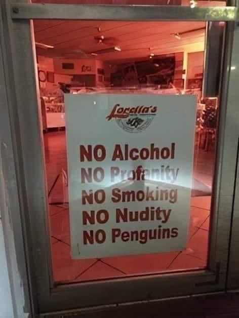 The image shows a white sign posted on the glass entrance door of Farella's restaurant. The sign lists standard, expected establishment rules in bold red text: **NO Alcohol**, **NO Profanity**, **NO Smoking**, **NO Nudity**, then abruptly ends with the completely unrelated and absurd rule **NO Penguins**. The humor comes from the jarring, nonsensical addition of a rule against penguins—an animal with no logical connection to typical restaurant policies—creating a wholesome, silly, unexpected joke.