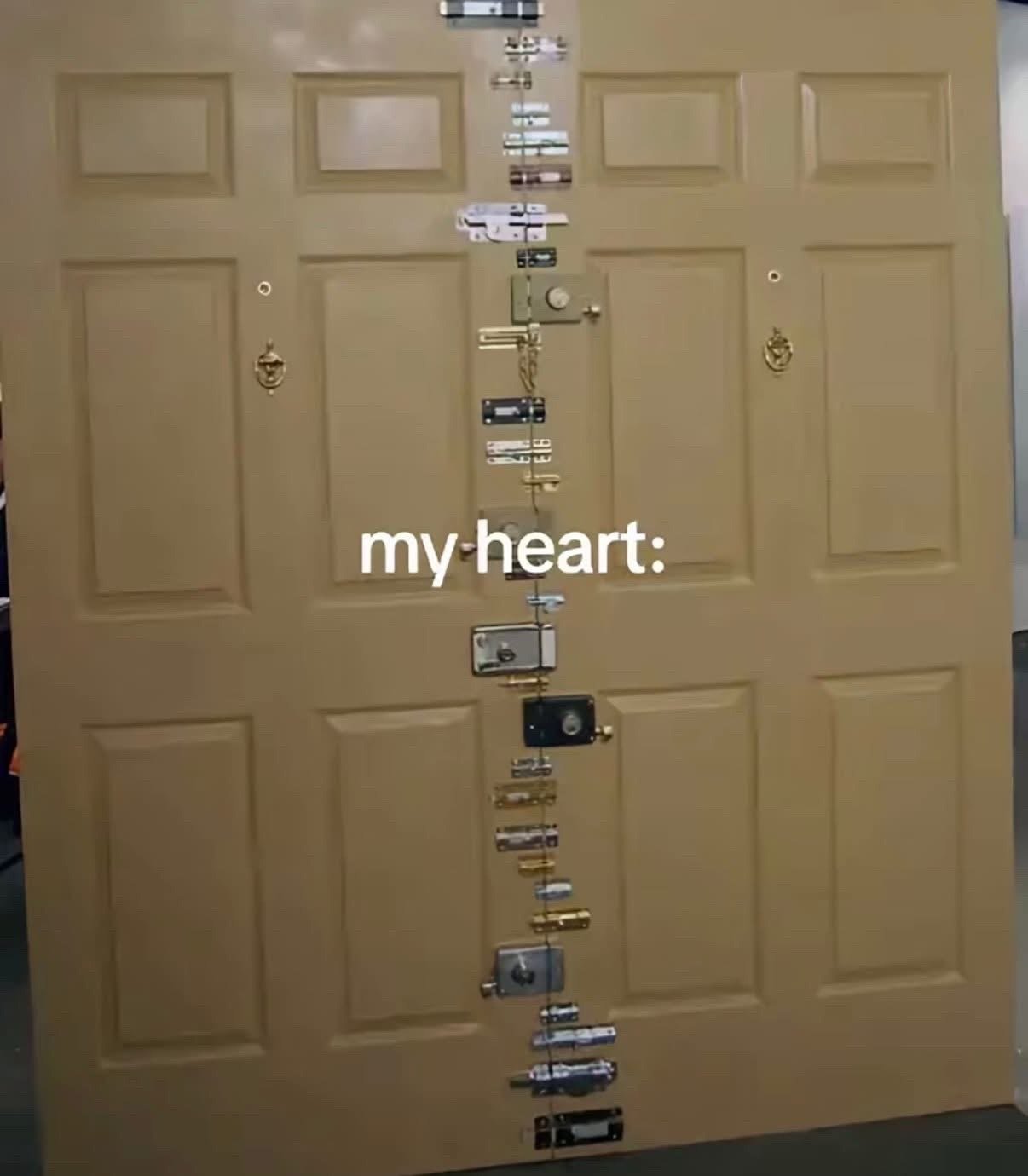 The image displays a beige double residential door fitted with an absurdly excessive number of varied locks, deadbolts, and latches stacked vertically all along the seam where the two doors meet, implying an extreme level of security and reluctance to open. White text overlaid on the center of the door reads "my heart:", using the over-secured door as a visual metaphor for being emotionally closed off, heavily guarded, and unwilling to let other people get close, usually as a result of past romantic or emotional hurt. It is a relatable humorous joke for people who struggle with trust issues in relationships.