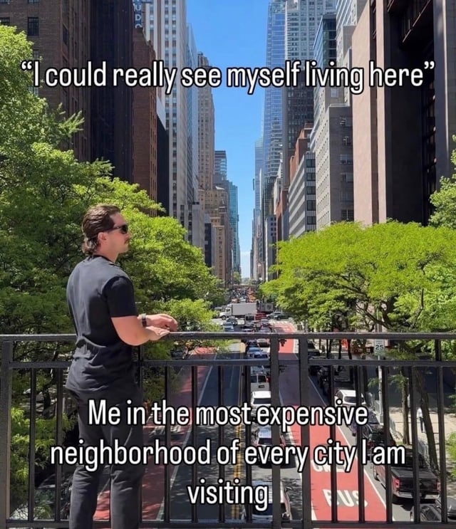 This meme features a photograph of a man wearing sunglasses and a black t-shirt, leaning on a metal railing to overlook a bustling, upscale city avenue lined with skyscrapers, green leafy trees, and busy traffic under a clear blue sky. There are two text overlays: the top line reads "I could really see myself living here", and the bottom line reads "Me in the most expensive neighborhood of every city I am visiting". The joke pokes fun at the common, relatable experience of admiring nice, high-cost areas while traveling, and briefly entertaining the unrealistic idea that one could afford to reside there, ignoring the exorbitant local cost of living.