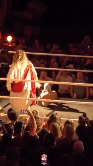 This photo captures an absurd, unexpected scene: a person dressed in the traditional popular depiction of Jesus Christ (a white robe with a red sash) stands inside a boxing/wrestling ring, with another person seated on the ring mat in front of him. A crowd of spectators surrounds the ring, many holding up cell phones to take photos of the strange scene. The humor comes from the juxtaposition of the religious figure of Jesus being placed in a combat sports fighting ring, creating a confusing and funny unexpected situation.