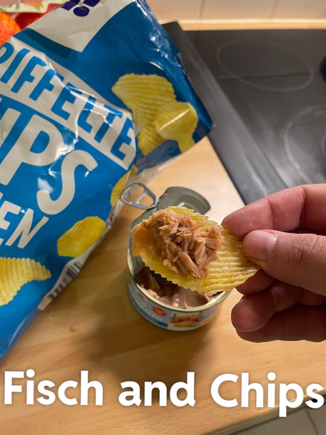 A close-up photograph depicting a wooden kitchen surface with a partially open blue bag of 'Piffelte Chips' (ridged potato chips) and an open can of tuna. A human hand is shown holding a single ridged chip topped with tuna from the can. At the bottom of the image, bold white text reads 'Fisch and Chips,' a pun combining the German word for fish ('Fisch') with 'Chips,' referencing the classic British dish 'Fish and Chips' but substituting traditional fried fish with canned tuna on potato chips.