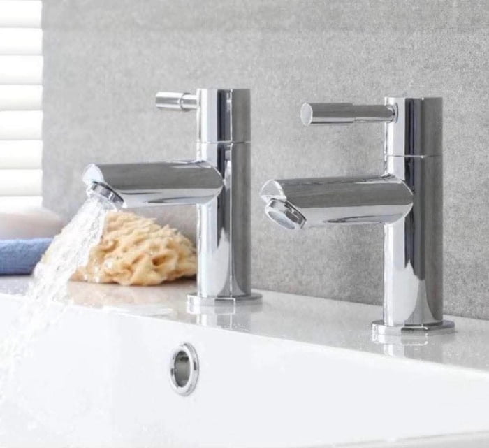 A bathroom sink counter featuring two chrome-plated faucets. The left faucet is actively running water, which flows into the white sink basin below, creating a small stream. The right faucet remains turned off, with no water flow. On the countertop between the faucets, there is a yellow, porous sponge and a folded blue towel. The background consists of a light gray textured wall, and a window with white horizontal blinds is partially visible on the left side, allowing soft light into the scene. The sink itself is white with a small overflow hole near the top edge.