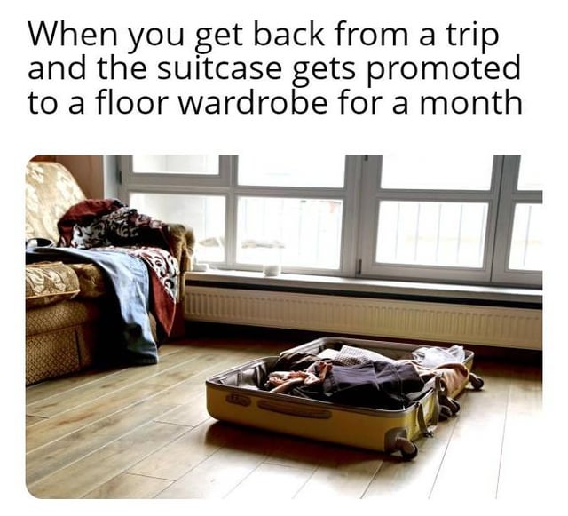 The meme features a top text caption paired with a real life photograph. The top text states 'When you get back from a trip and the suitcase gets promoted to a floor wardrobe for a month'. The image below displays an open yellow suitcase full of clothes left unpacked on the wooden floor of a living room, next to a patterned couch with additional clothes draped over it, near large windows. The meme jokes about the common relatable habit of procrastinating unpacking after travel, leaving the suitcase open on the floor to use as a makeshift wardrobe for weeks instead of putting clothes away in a proper closet.