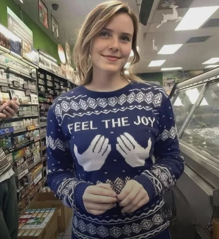A photograph of a person standing in a retail store, wearing a blue Christmas sweater adorned with white festive patterns. The sweater prominently features the text 'FEEL THE JOY' in white capital letters, with two white hand designs positioned below the text. The individual has a smiling expression and stands with hands clasped in front. The background shows store shelves stocked with various products and a refrigerated display case, indicating a typical retail environment. The image conveys a lighthearted, festive atmosphere associated with Christmas celebrations.