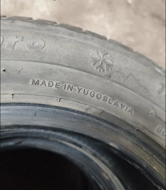 A close-up photograph of a tire's sidewall, focusing on the embossed text 'MADE IN YUGOSLAVIA'. The tire appears aged with visible wear, including slight cracking and discoloration. A snowflake symbol is present near the text, suggesting it may be a winter tire. The background is out of focus, emphasizing the tire's details and the historical reference to Yugoslavia.