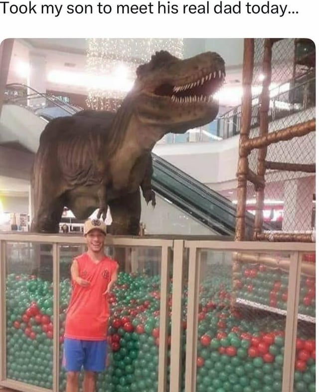 A humorous meme image featuring a man in a red shirt, blue shorts, and a beige cap standing in front of a ball pit filled with green and red balls in a mall environment. Behind him is a large, realistic Tyrannosaurus rex statue with its mouth agape, displaying sharp teeth and a fierce expression. The statue is positioned above the ball pit, with a mall escalator visible in the background. The top text of the meme reads, "Took my son to meet his real dad today...", creating a comedic absurdity by jokingly implying the dinosaur statue is the child's biological father.