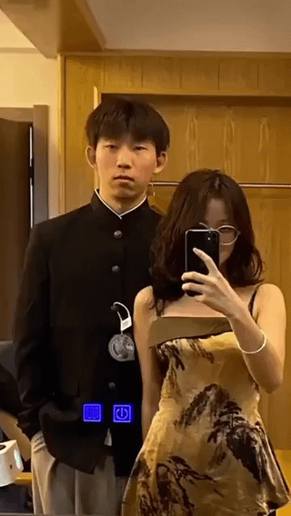 This image captures a couple taking an indoor mirror selfie. The woman on the right wears a strapless landscape-print dress and glasses, holding a smartphone to snap the photo with a neutral, focused expression. Behind her, the man wears a black button-up jacket with a large padlock attached to the front, standing with hands in his pockets and a deadpan, slightly annoyed expression directed at the camera. Edited blue UI-style icons (resembling media controls) are overlaid on the lower part of his jacket, adding a comedic, video-screenshot effect. The gag plays on the humorous implication of being "locked into" a relationship, using the padlock as a silly, exaggerated accessory. The background features wooden cabinetry, indicating a home setting.
