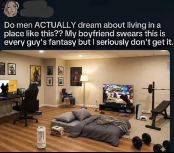 The image features a photograph of a casual, lived-in room with elements commonly associated with a young man's living space. Visible items include a gaming setup (monitor, black chair, desk), a bed with gray bedding, a TV displaying a video game, gym equipment (barbell, dumbbells, weight bench), multiple posters on the walls, and a Monster Energy drink can on the wooden floor. Overlaid text at the top poses a question: 'Do men ACTUALLY dream about living in a place like this?? My boyfriend swears this is every guy's fantasy but I seriously don't get it.' The room has a relaxed, personal atmosphere with scattered items suggesting daily use.
