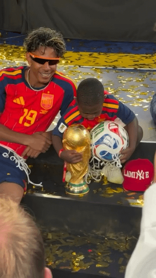 The photograph captures Spanish professional football player Lamine Yamal, wearing his Spain national team #19 jersey and orange-trimmed novelty sunglasses, smiling as he sits on a stage covered in golden confetti during a championship victory celebration. Seated directly next to him is his young younger brother, who holds the iconic golden FIFA World Cup trophy in one hand and an official match ball in the other, leaning over the items and drawing focus away from the star player. The meme is captioned 'When the side quest becomes the main character', referring to how the small child with the coveted trophy is the unexpected center of attention in the shot, overshadowing the famous athlete beside him.