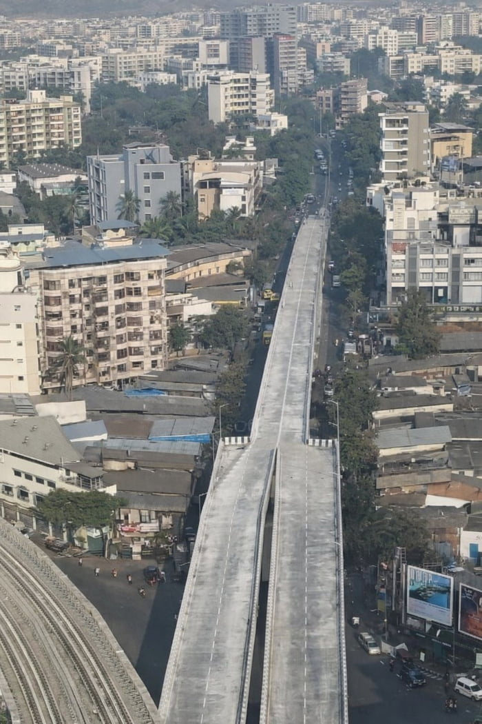 An aerial photograph of a newly constructed elevated bridge in Mumbai, India. The bridge initially spans 4 lanes (two in each direction) but abruptly narrows to 2 lanes ahead, creating a potential traffic bottleneck. Surrounding the bridge is dense urban development, including high-rise buildings, low-rise structures, trees, and a busy street below with visible vehicles and pedestrians. The image highlights a perceived infrastructure planning issue where a wider bridge unexpectedly reduces in capacity.