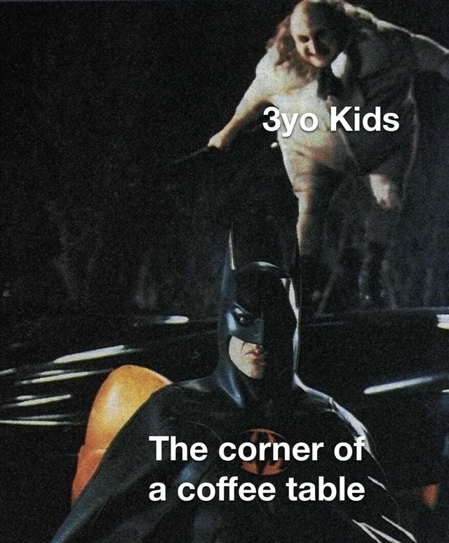 The meme features a screenshot from a Batman film, showing Batman in his iconic black costume with a serious expression, standing near the Batmobile. Above him, a character (likely Catwoman from the original scene) is labeled "3yo Kids" and appears to be leaning down toward Batman with a mischievous or determined posture. Below Batman, the text reads "The corner of a coffee table." The humor lies in the analogy that 3-year-old children are irresistibly drawn to coffee table corners (often resulting in bumps), similar to how the character is visually drawn to Batman, playing on the "like a moth to a flame" trope.