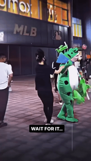 An outdoor nighttime scene depicting a person in dark clothing interacting with a green dragon mascot. The mascot features a blue triangular element on its head and green appendages. In the background, illuminated signs for 'IMAX' and 'MLB' are visible, indicating a commercial or entertainment district. Other people are present in the background, suggesting a public area. A prominent text overlay at the bottom reads 'WAIT FOR IT...', creating a sense of anticipation for an upcoming action or event involving the mascot and the person.