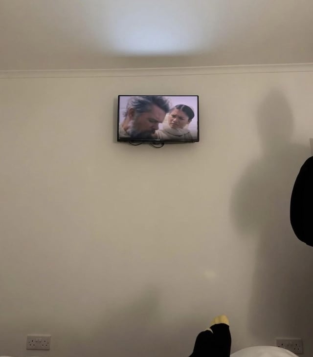 The image is shot from a first-person perspective of someone lying down in a room, watching a film on a very small television mounted extremely high on a plain white wall near the ceiling. The film playing on the tiny screen is Christopher Nolan's acclaimed work *Oppenheimer*, with two lead characters visible on the display. A portion of the viewer's foot wearing a black sock with yellow accents is visible at the bottom right of the frame, emphasizing how far the viewer is from the inconveniently placed screen. The core joke is sarcastic: the poster refers to the highly praised film as Nolan's masterpiece, but the terrible, low-quality viewing setup completely undermines the experience of engaging with the respected work.