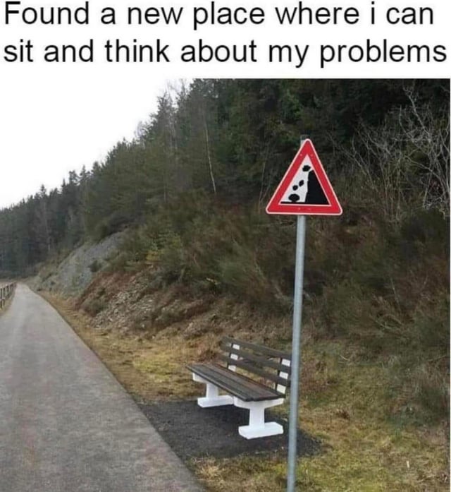 A meme image depicting a scenic outdoor setting with a paved road running alongside a forested hillside. A wooden bench with white legs is positioned near the road, seemingly offering a peaceful spot to sit. Adjacent to the bench stands a triangular road sign with a red border, warning of falling rocks (depicted by black rock icons on a white background). At the top of the image, overlaid text reads: 'Found a new place where i can sit and think about my problems.' The humor stems from the irony of seeking a relaxing, problem-solving spot being juxtaposed with a warning sign indicating potential danger from falling rocks, undermining the intended relaxation.