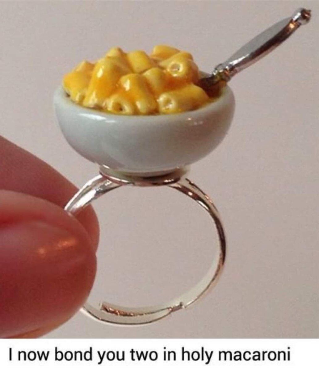 The image shows a tiny, handmade ring designed to look like a small ceramic bowl filled with macaroni and cheese, complete with a tiny spoon resting inside the bowl. A person's fingers are holding the ring to display it. Below the ring, there is text that reads: "I now bond you two in holy macaroni" — this is a playful pun on the traditional wedding phrase "holy matrimony," replacing it with "macaroni" to tie into the ring's food-themed design. The meme humorously frames this whimsical food ring as an alternative to a wedding or engagement ring, combining the idea of marriage bonds with a love for comfort food, creating a lighthearted, wholesome joke.