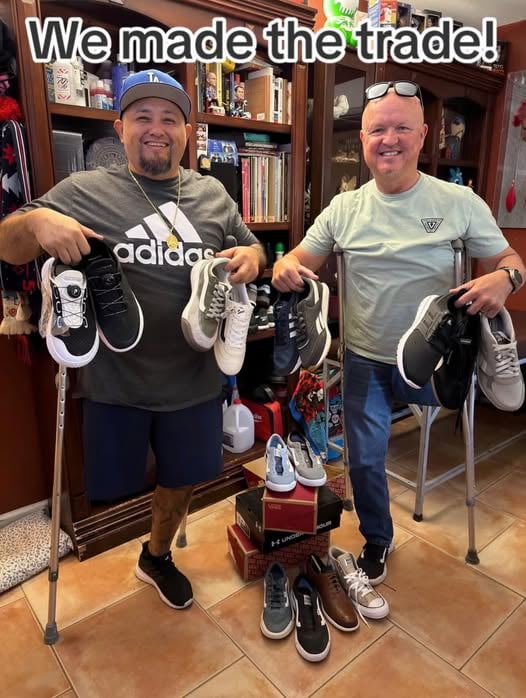 Two men, both using crutches, stand indoors in a room filled with shelves of books, shoes, and various items. The man on the left wears a gray Adidas t-shirt, blue shorts, and has a prosthetic leg; he holds two pairs of sneakers (black and white, and gray and white). The man on the right wears a light green t-shirt and jeans, holding two pairs of shoes (black and gray). Between them are stacked shoe boxes with additional shoes on top. Both men appear happy and proud, with the text 'We made the trade!' prominently displayed at the top of the image, indicating a successful shoe exchange. The atmosphere is warm and wholesome, highlighting positivity and community.