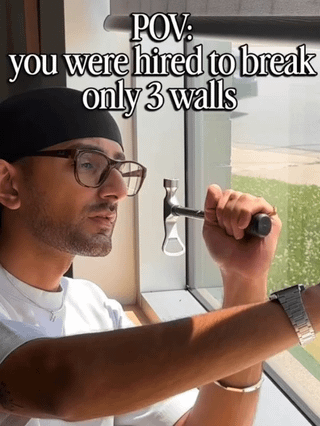 This is a POV-style comedy meme. It depicts a man wearing a black beanie, thick-rimmed glasses, and a white t-shirt holding a small hammer up to an interior glass window as if he is about to hit it. Overlaid text at the top reads "POV: you were hired to break only 3 walls". The humor comes from a double pun: the man has already completed his assigned task of breaking 3 structural walls, and the next surface in front of him is effectively the "fourth wall", referencing both the physical fourth wall of the space (the glass window) and the theatrical/media term "breaking the fourth wall", where a work of fiction addresses the audience directly. The man's slightly conflicted expression implies he realizes he is about to go beyond the scope of the work he was hired to do.