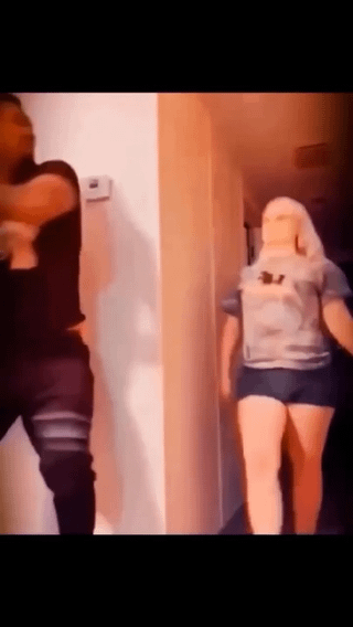 This is a low-resolution, warm orange-tinted still frame from a short video clip. The setup implies a man on the left side of the frame is recording 'wholesome couple content' of his partner walking towards him down a residential home hallway. The punchline revealed on closer inspection (the 'zoom in' referenced in the title) is that the figure approaching him is not a real woman, but a life-sized sex doll, turning the expected cute romantic couple premise into a crude, unexpected humorous twist.