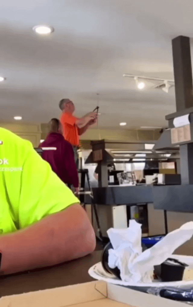 A photograph of a workplace setting, possibly an office or restaurant kitchen. In the foreground, a person wearing a bright neon yellow shirt has their forearm resting on a table, partially obscuring the view. In the background, two individuals are visible: one person in an orange shirt standing on a ladder, using a tool to reach up towards the ceiling (likely fixing a light fixture or ceiling component), and another person in a maroon shirt standing beside the ladder, observing or assisting. The environment features multiple ceiling lights, a ventilation hood, and various office or kitchen equipment, suggesting a casual work environment.